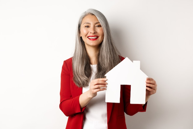 Asian female real estate agent showing paper house cutout, broker smiling friendly and selling property, standing over white wall.