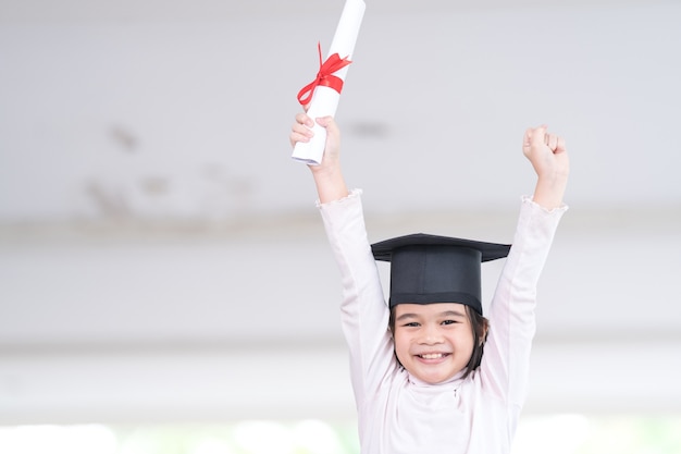 Asian female school kid graduate in a graduation cap holds a rolled certificate paper