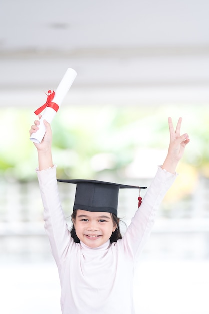 Asian female school kid graduate in a graduation cap holds a rolled certificate paper