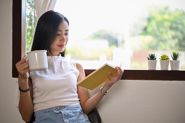 窓の近くに座ってコーヒーを飲みながらお気に入りの本を読むアジアの女性