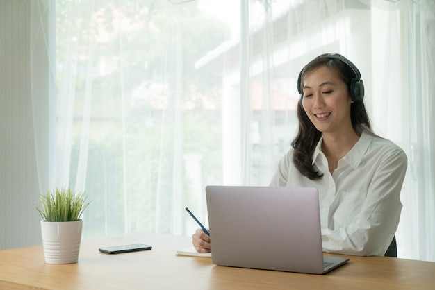 Femmina asiatica sorridente e videochiamata in linea dal computer portatile notebook waring cuffie wireless e prendere appunti alla scrivania.