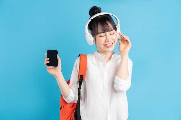 Asian female student wearing backpack behind her back, holding smartphone, neck wearing wireless earphones, holding paper cup