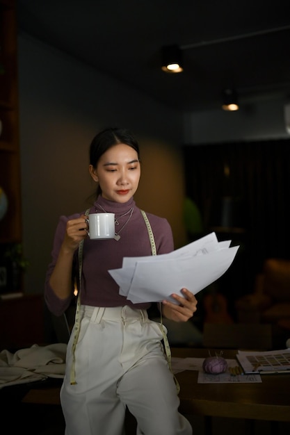 An Asian female tailor focuses on checking her model sketches while sipping coffee in a studio