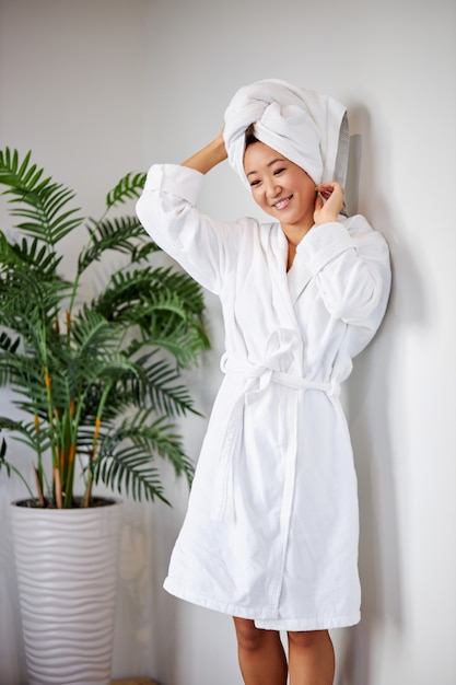 Фото Азиатская женщина трогает голову в полотенце, стоит после душа, наслаждается временем для себя, концепция красоты