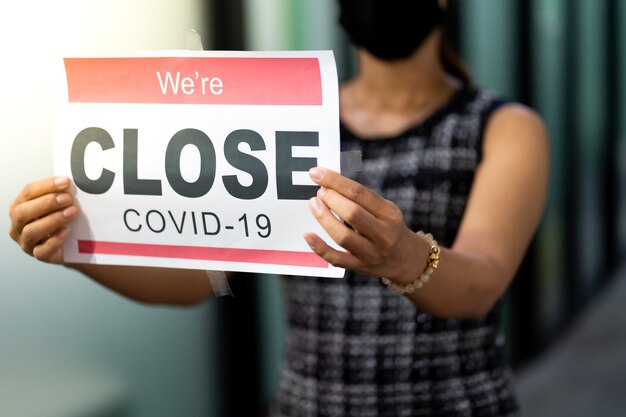 Asian female wearing medical mask puts a Temporary closed due COVID-19 pandemic sign banner on door and windows at office