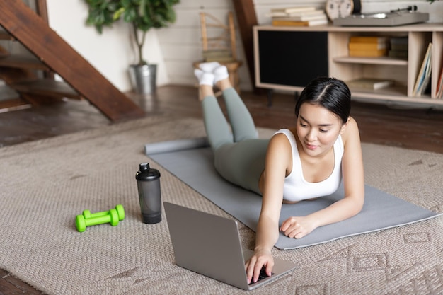 Asian fitness girl taking online fitness course watching sports video exercising at home on floor ma...