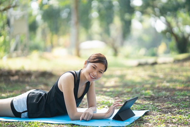 ビデオクリップによるとヨガをしているアジアのフィットネス女性。タブレットを使用して公園でヨガをしているアジアの女性