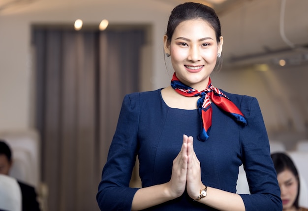 Asian flight attendant posing with smile at middle of the aisle inside aircraft passenger seat on the background