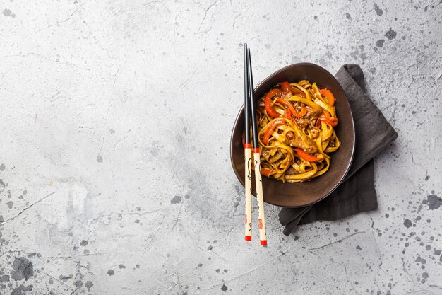 Asian food, stir fry udon noodles with meat and vegetables in a bowl on gray stone surface, top view