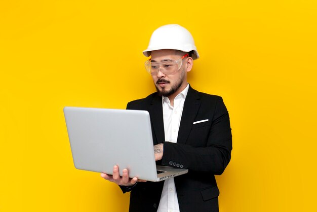 Asian foreman in suit uses laptop on yellow isolated background korean civil engineer in hard hat