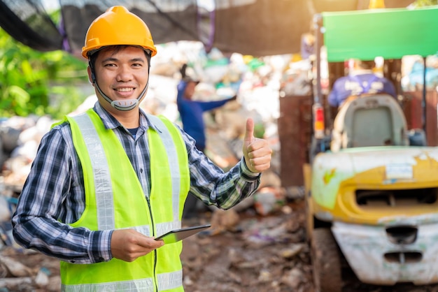 Asian foreman thumbs up and holding tablet and looking at recycle waste factory with a forklift on the background