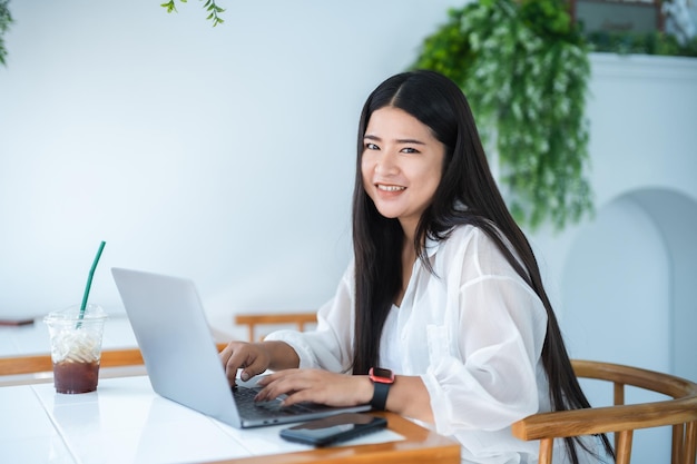 Donna d'affari freelance asiatica che indossa smartwatch parlando facendo utilizzando il lavoro casual con il computer portatile per navigare in internet, chattare e bloggare e smartphonenotebook in caffetteria