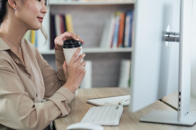 Donna asiatica freelance sorridente tenendo una tazza di caffè caldo e lavorando su un computer portatile su un tavolo di legno a casa imprenditrice donna che lavora per la sua attività a casa concetto di lavoro a casa