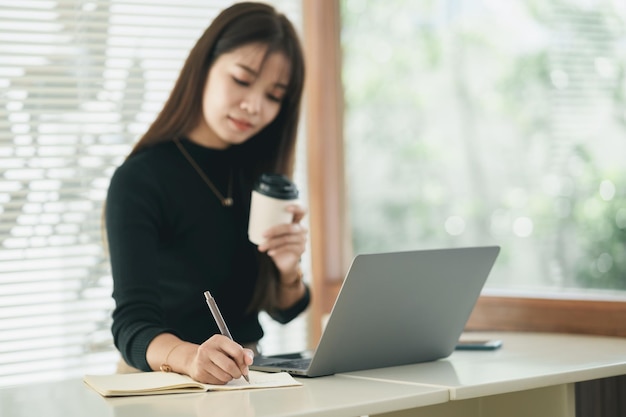 Donna asiatica freelance che sorride usando la penna che fa gli avvisi il suo taccuino e lavora al computer portatile sul tavolo a casa imprenditrice che lavora per la sua attività al bar lavoro d'affari al concetto di caffetteria