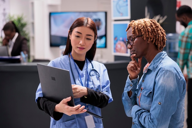 Asian general practitioner using computer to give care instructions to young african american woman in medical clinic living room. Female patient listening attentively to doctor recommendations.