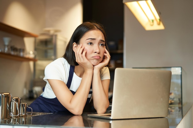 Asian girl barista standing behind counter in cafe looking as at laptop working while feeling depres