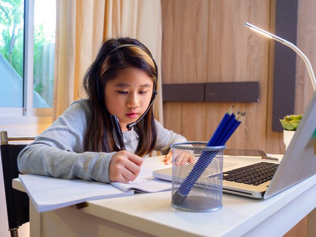 Asian girl in headphones sit at desk study online on laptop Kid wear headset handwriting in notebook