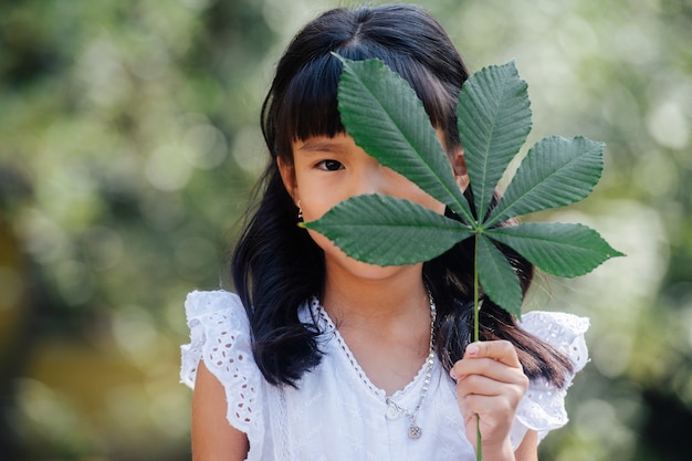 彼女の顔の上に6枚の葉で茎を保持しているアジアの女の子。肖像画。彼女は公園に立っていて、背景がぼやけています。