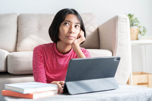 Asian girl is sitting thinking and reviewing lessons at tablet with hand on face in the living room