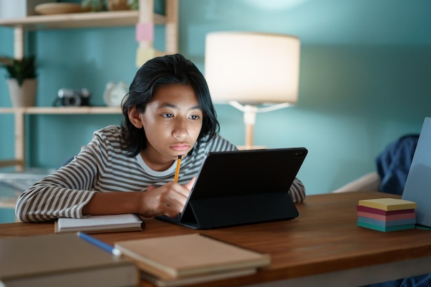 写真 アジアの女の子は、自宅の夜に机の上に座っている間、タブレットでインターネットを介してオンラインで勉強しています。自宅でのオンライン学習の概念