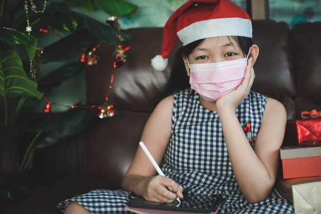 An asian girl is thinking of good wishes to write for others. a girl wearing a mask uses a tablet to write during christmas and new year's