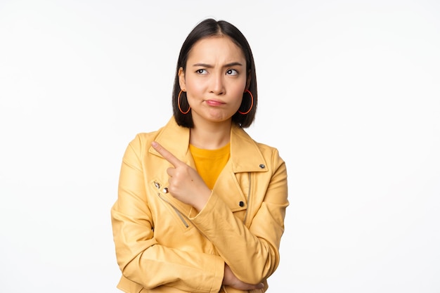 Asian girl looking skeptical pointing finger left at smth strange staring doubtful and displeased at product banner standing against white background