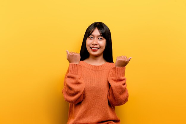 Ragazza asiatica bella che indossa un abito casual arancione sfondo giallo festeggia la vittoria con un sorriso felice e l'espressione del vincitore con un'espressione felice della mano alzata