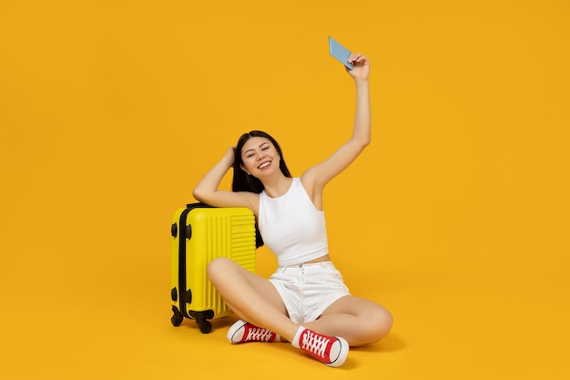 An asian girl sits near a suitcase on a yellow background