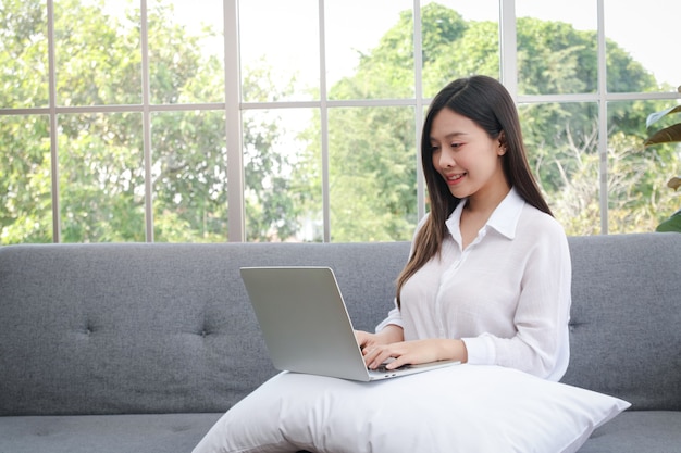 Asian girl sitting on the sofa in the living room at home Working on a laptop computer. Work from home concept, social distancing During the COVID-19 pandemic