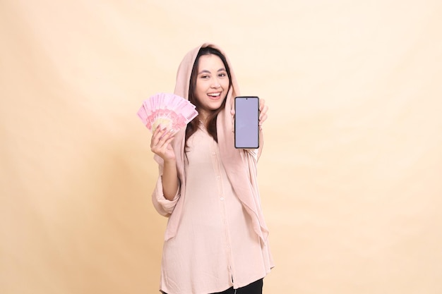 Photo asian girl smiling happily at the camera carrying rupiah money and holding the cellphone gadget scre