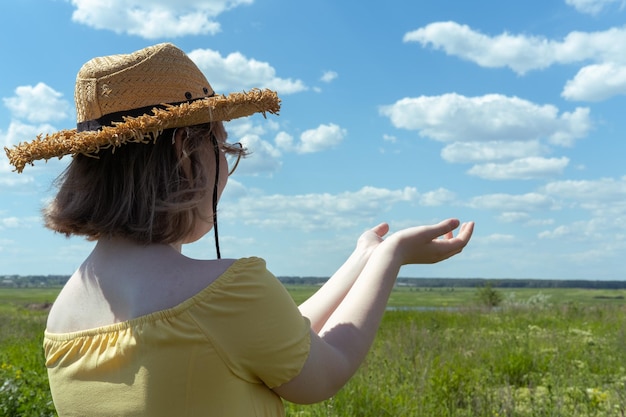 Ragazza asiatica in un cappello di paglia e un vestito giallo tiene insieme i palmi delle mani contro il cielo blu con nuvole