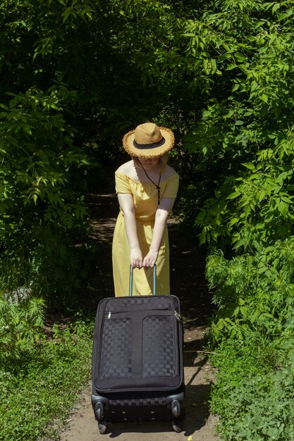 Asian girl in straw hat and yellow dress with big black suitcase among green trees Summer vacation