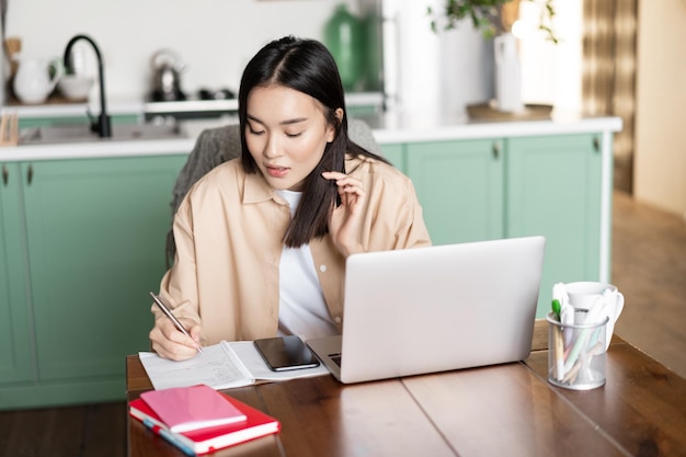 Asian girl student doing homework at home young woman taking notes working on laptop and writing dow...