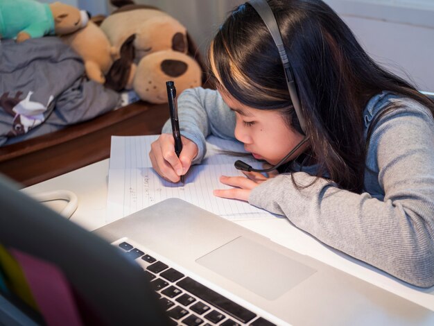 アジアの女の子がラップトップでオンラインで勉強する子供はヘッドセットを着用してノートブックで書く検疫でインターネットレッスンを使用する学生は学校のインターネットオンラインクラスで学ぶCovid19