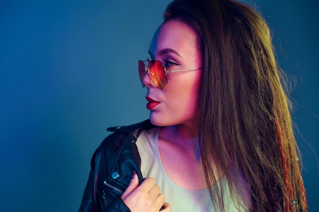 Asian girl in sunglasses and black leather jacket posing against dark blue background