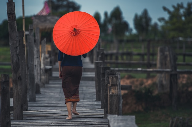 Asian girl on traditional clothes with a red umbrella crossing wooden bridge