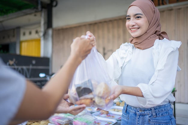 An asian girl in a veil gives takjil food orders in plastic bags to buyer