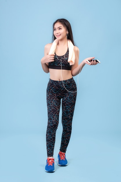 An Asian girl wearing a leggings and plugging in a phone earphone to exercise on a pastel blue studio scene.