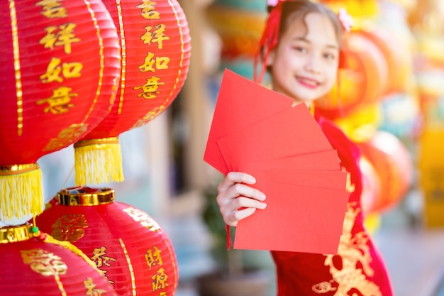 Ragazza asiatica che indossa cheongsam rosso cinese tradizionale, con buste rosse in mano e lanterne con scritto sopra il testo cinese benedizioni è una benedizione della fortuna per il capodanno cinese