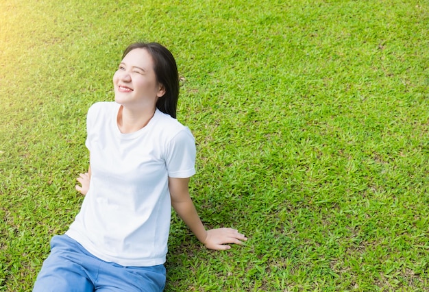 Asian girl in white shirt Come out to relax and enjoy the warm sunlight in the evening On a happy rest day from work Outside sitting on the green lawn looking up at the light and smiling in the park