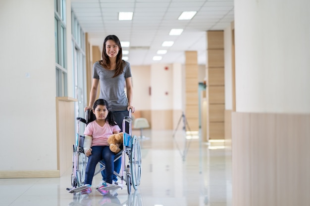 Asian girl with a broken arm wearing a cast setting on wheelchair with her mother at hospital