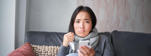 asian girl with influenza sits at home cries and looks upset holds smartphone feels unwell and