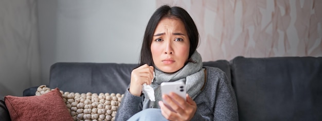 asian girl with influenza sits at home cries and looks upset holds smartphone feels unwell and