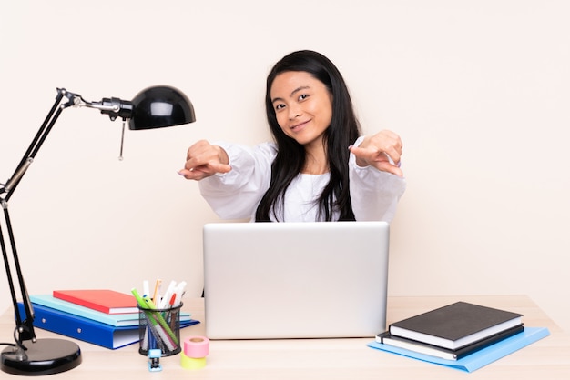 Asian girl in a workplace with a laptop on beige wall points finger at you while smiling