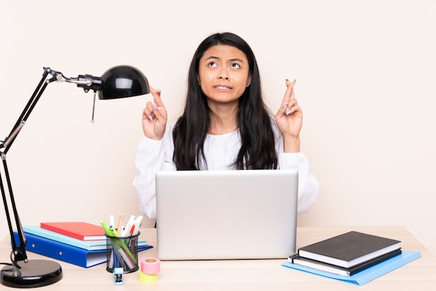 Asian girl in a workplace with a laptop on beige wall with fingers crossing and wishing the best