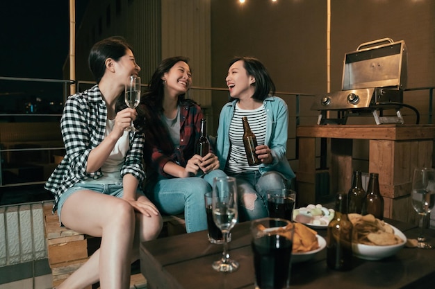 옥상 파티에서 웃고 있는 아시아 소녀들. 밤에 야외에 앉아 술을 마시며 수다를 떨고 있는 젊은 여성. 맥주와 와인과 함께하는 바베큐 이벤트.