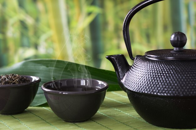 Asian green tea set with black china kettle on bamboo mat with dried green tea in bowl. 