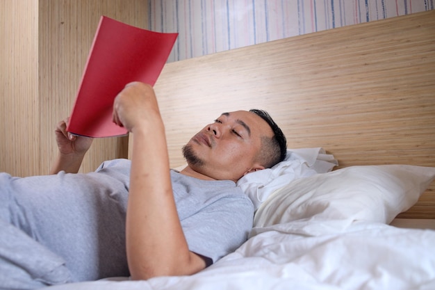 Asian Guy reading book lying in comfortable bed in bedroom at home