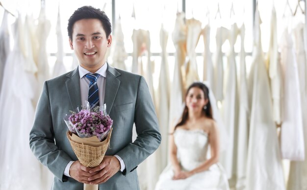 Asian handsome groom in grey formal suit with necktie standing look at camera with bouque and beautiful happy bride in white long wedding dress in blur background.