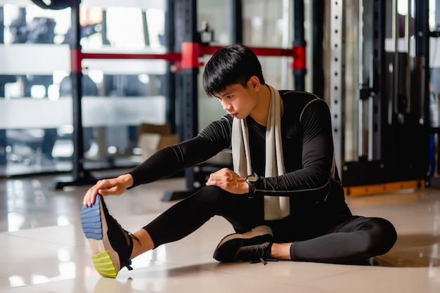 Un bell'uomo asiatico che indossa abbigliamento sportivo e smartwatch seduto sul pavimento e allunga i muscoli delle gambe prima dell'allenamento in palestra,